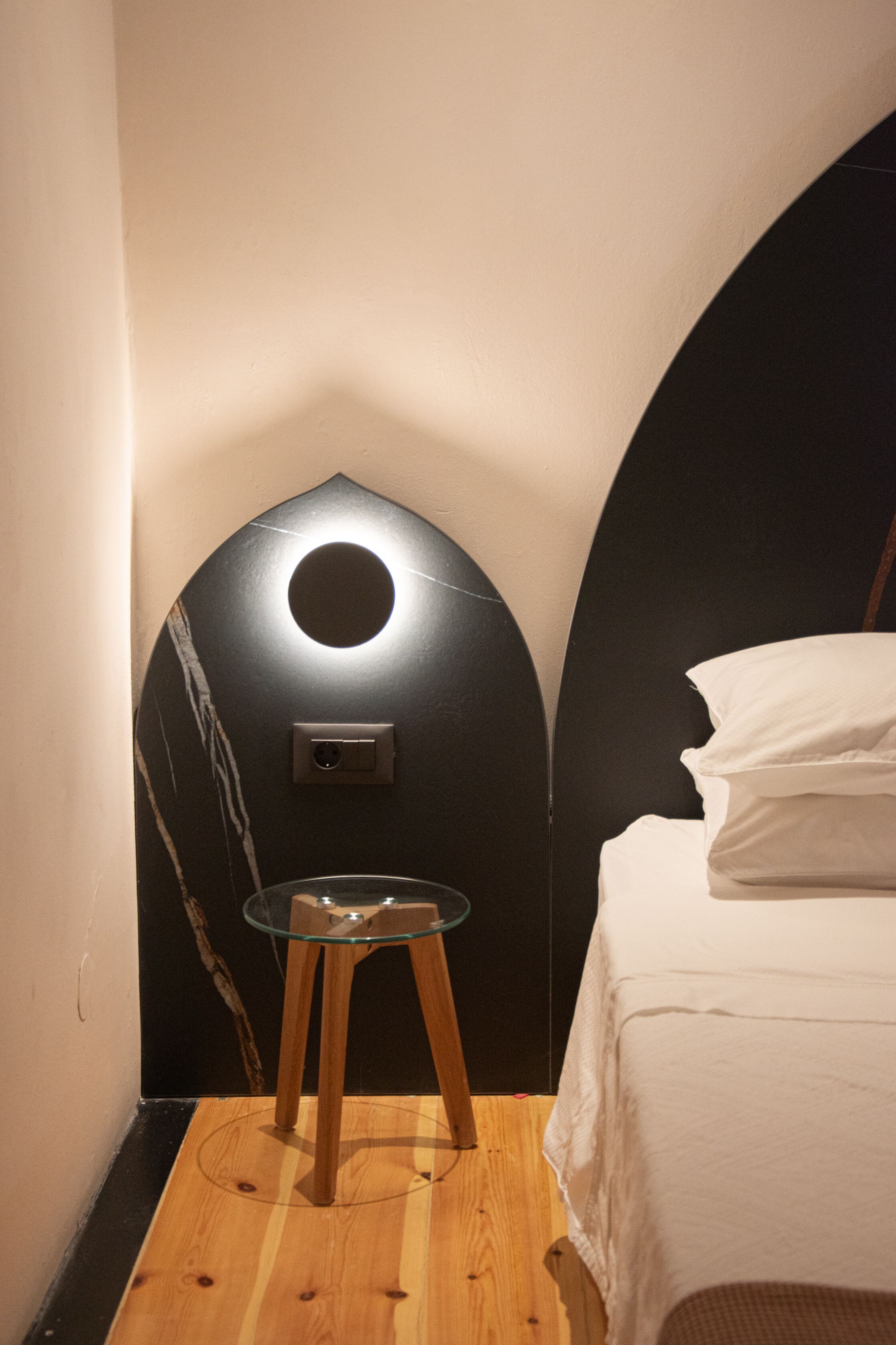 Quarto com cama, uma pequena mesa de vidro e luminária na parede.