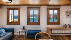 A room with three large windows offering views of the sea and a lighthouse. There's a blue sofa, a small table with a decorative item, and a bench with books. - San Salvatore Monastery (Chania)