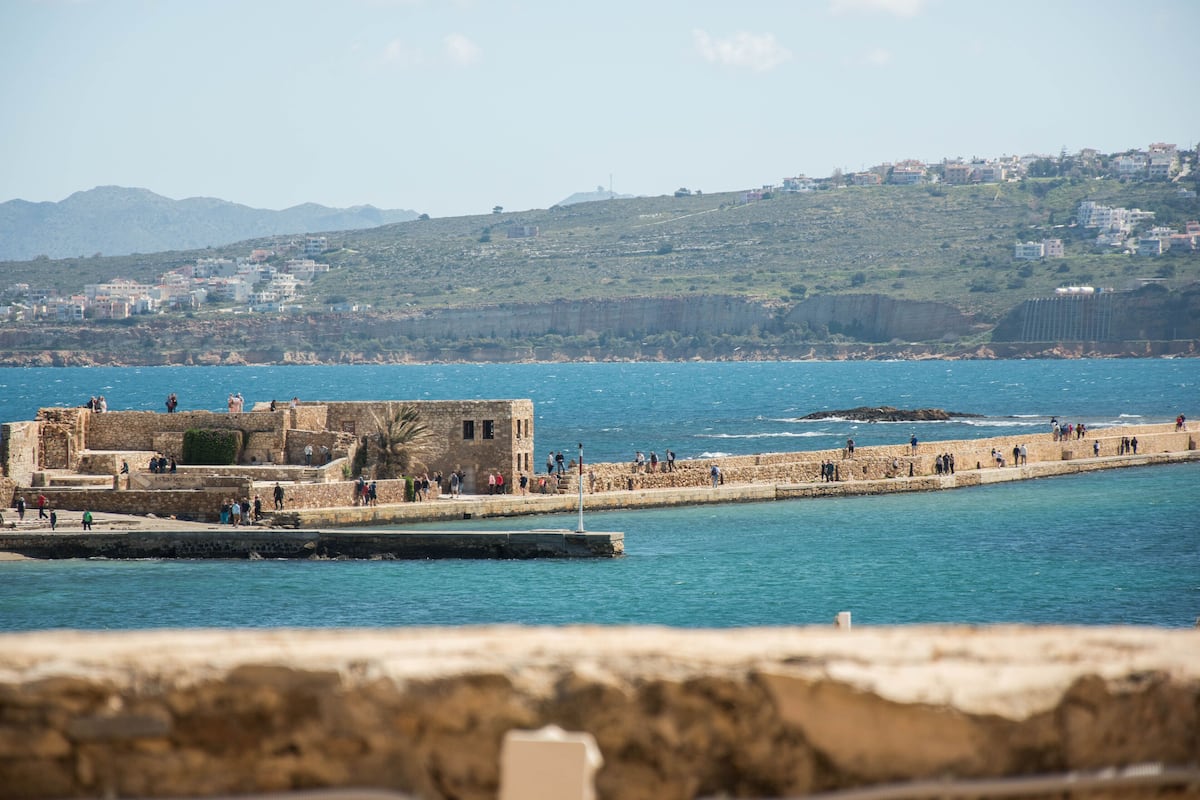 A coastal fortification with a stone wall and a tower, overlooking a calm sea and a hillside with buildings.