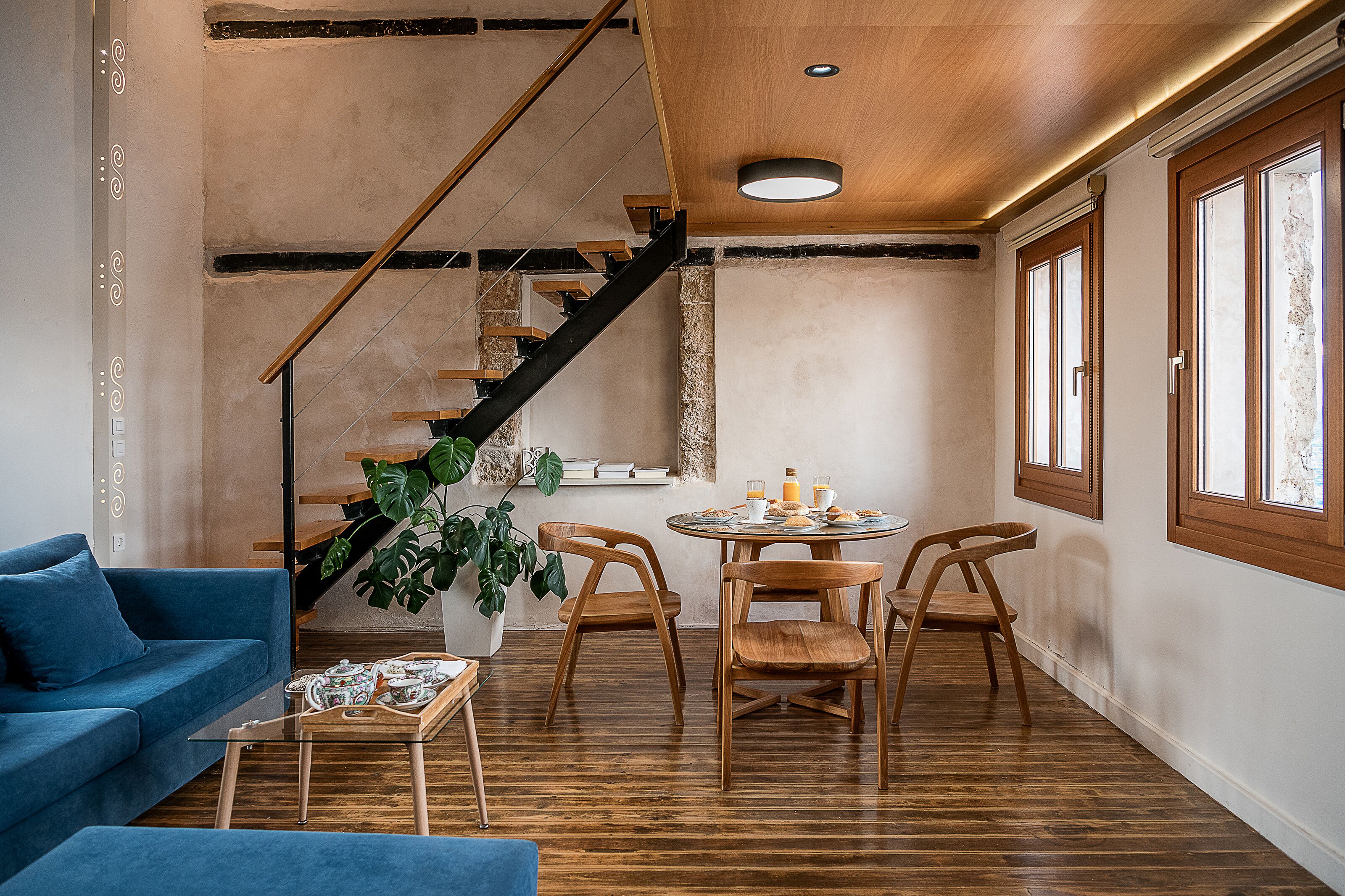 A modern living room with a blue sofa, wooden dining table, and chairs, a staircase, and a large window.