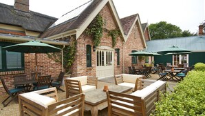 An outdoor patio with wooden furniture and green umbrellas, a brick building with a white door, and a green roof. - The Museum Inn (Blandford Forum)
