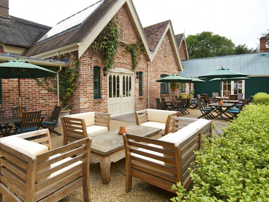 An outdoor patio with wooden furniture and green umbrellas, a brick building with a white door, and a green roof.