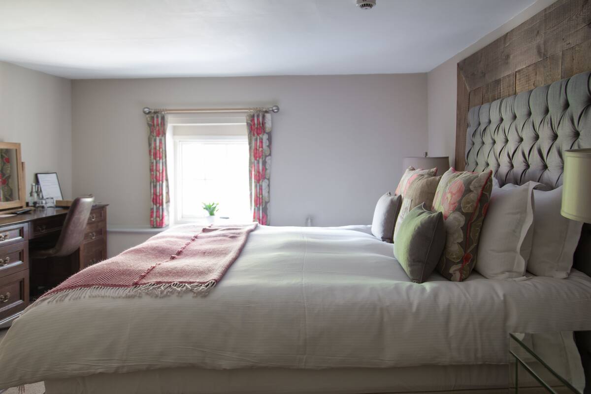 A bedroom with a large bed, a wooden headboard, a desk with a chair, and a window with curtains.