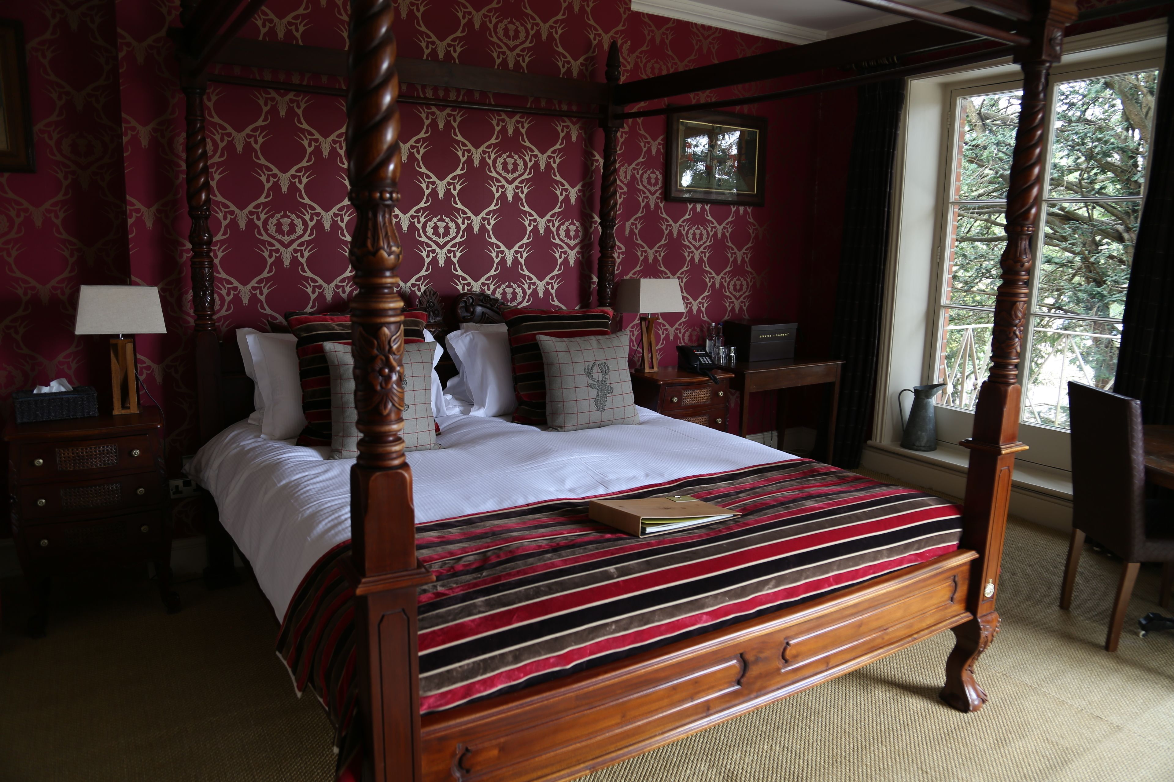 A four-poster bed with a striped bedspread, a wooden nightstand with a lamp, a chair, and a window with a view of trees.