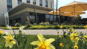 A modern building with a stone facade and large windows. There are yellow daffodils in the foreground and a yellow umbrella in the background. - Hotel Susato (Soest)