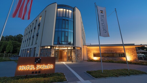 A modern hotel with a curved glass facade, a well-lit entrance, and a sign reading "HOTEL SUSATO". - Hotel Susato (Soest)