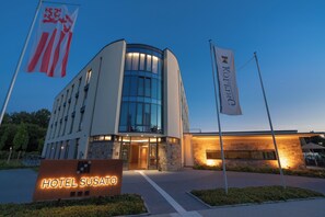 Un hotel moderno con una facciata in vetro curvo, un ingresso ben illuminato e un cartello con la scritta "HOTEL SUSATO".