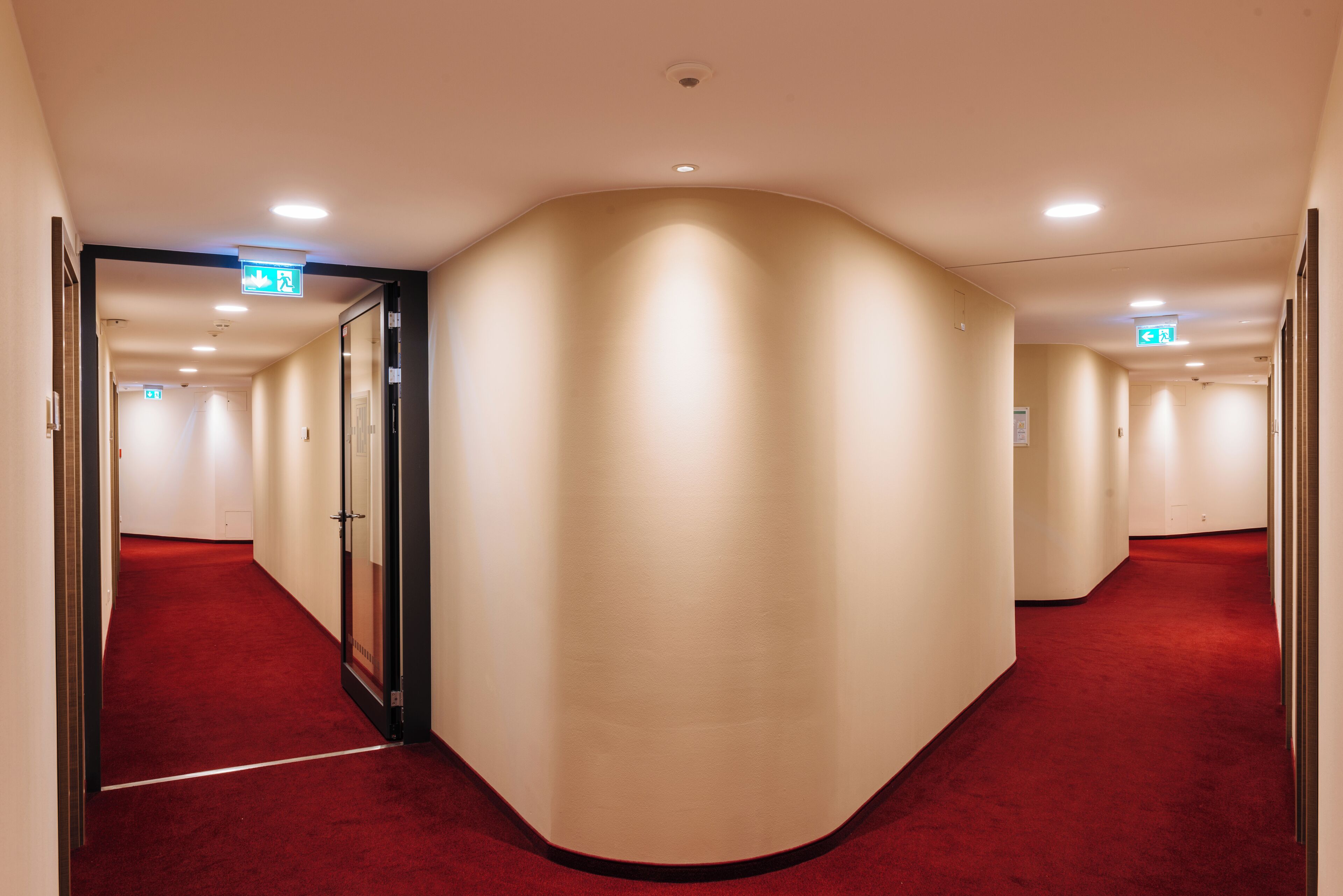 A hotel corridor with a red carpet, beige walls, and multiple exit signs.