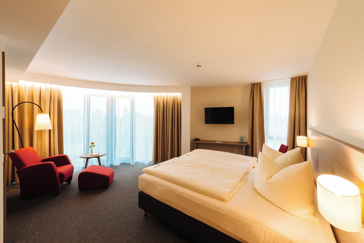 Ein Hotelzimmer mit einem großen Bett, einem roten Sessel, einem kleinen Tisch mit einer Lampe und einem Fernseher an der Wand.
