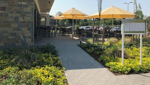 An outdoor cafe area with tables and chairs under yellow umbrellas, a stone building with a sign, and a parking lot in the background. - Hotel Susato (Soest)