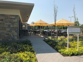 Un área de café al aire libre con mesas y sillas bajo sombrillas amarillas, un edificio de piedra con un letrero y un estacionamiento al fondo.