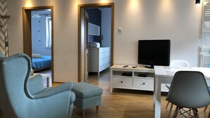 A modern living room with a sofa, a TV, a dining table, and a kitchen area visible through an open door. - Apartamenty Usteckie - Na Wydmie (Ustka)