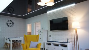 A modern living room with a yellow armchair, a white coffee table, a flat-screen TV mounted on a brick wall, and two wall clocks. - Apartamenty Usteckie - Na Wydmie (Ustka)