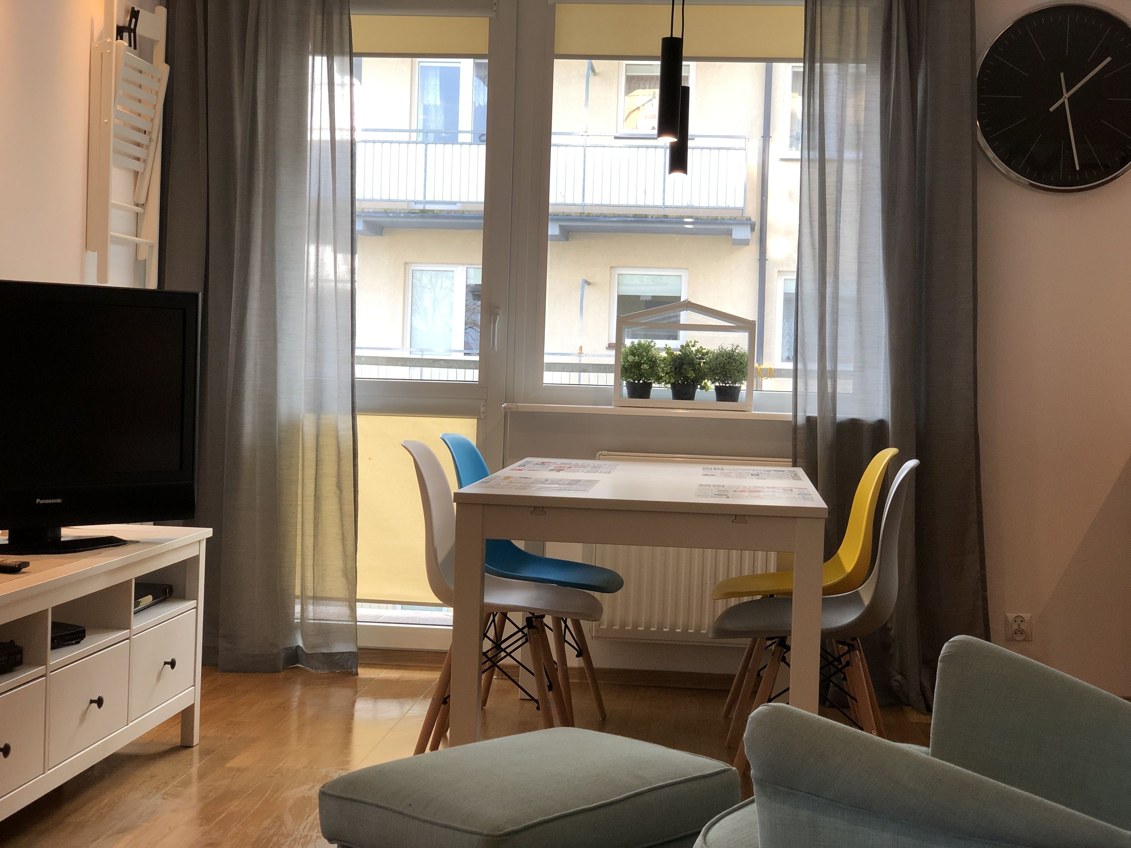 A modern living room with a flat-screen TV, a dining table with chairs, a sofa, and a view of a balcony with plants.