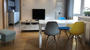 A modern living room with a white TV stand, a dining table with chairs, and a window with curtains. - Apartamenty Usteckie - Na Wydmie (Ustka)