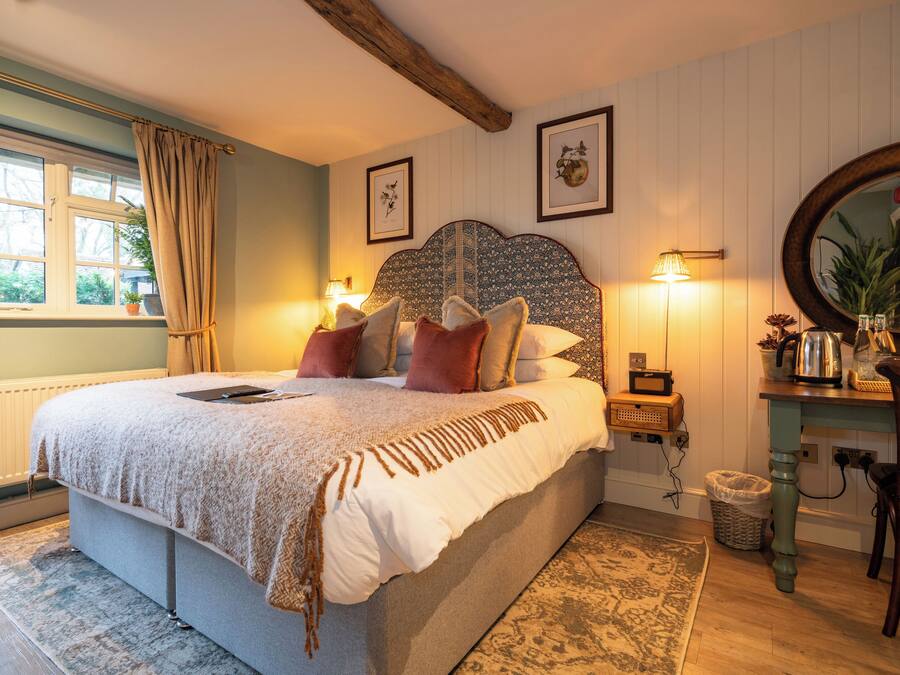 A bedroom with a large bed, a wooden headboard, a bedside table with a lamp, a mirror, and a view of the outdoors through a window.