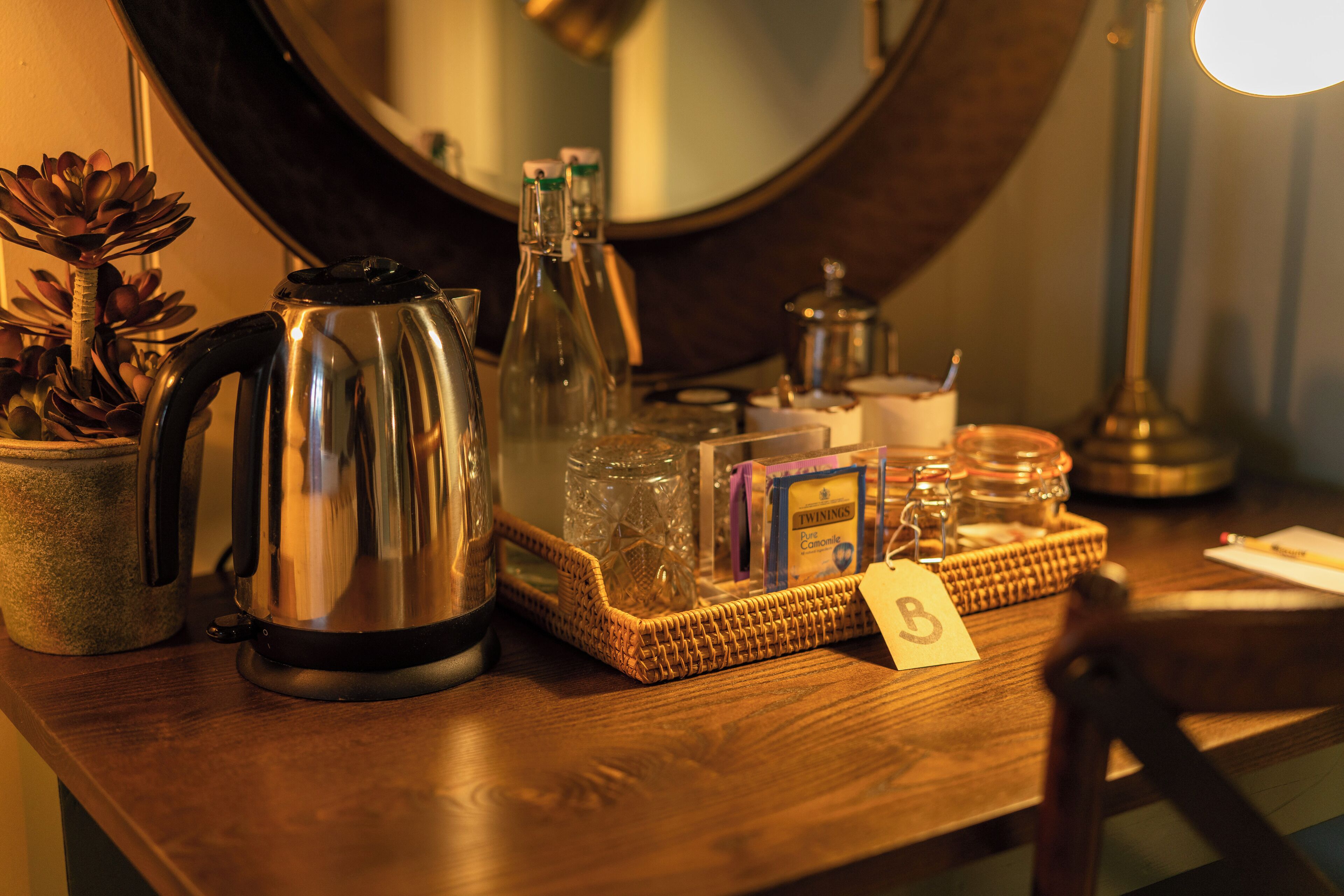 A wooden table with a stainless steel kettle, glass bottles, a woven basket, and a box of Twinings tea.