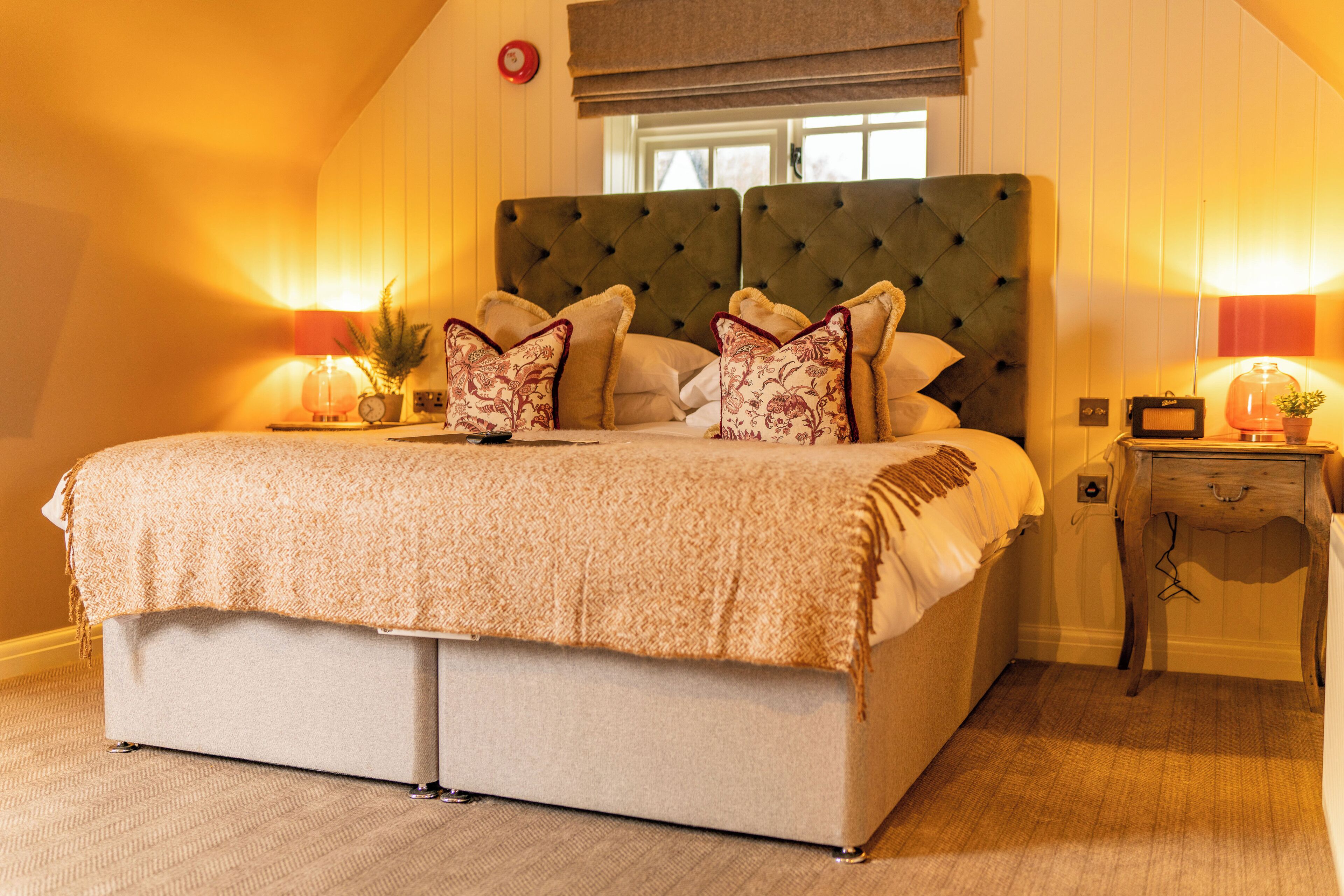A large bed with a tufted headboard, a bedside table with a lamp, and a window with a view of the outside.