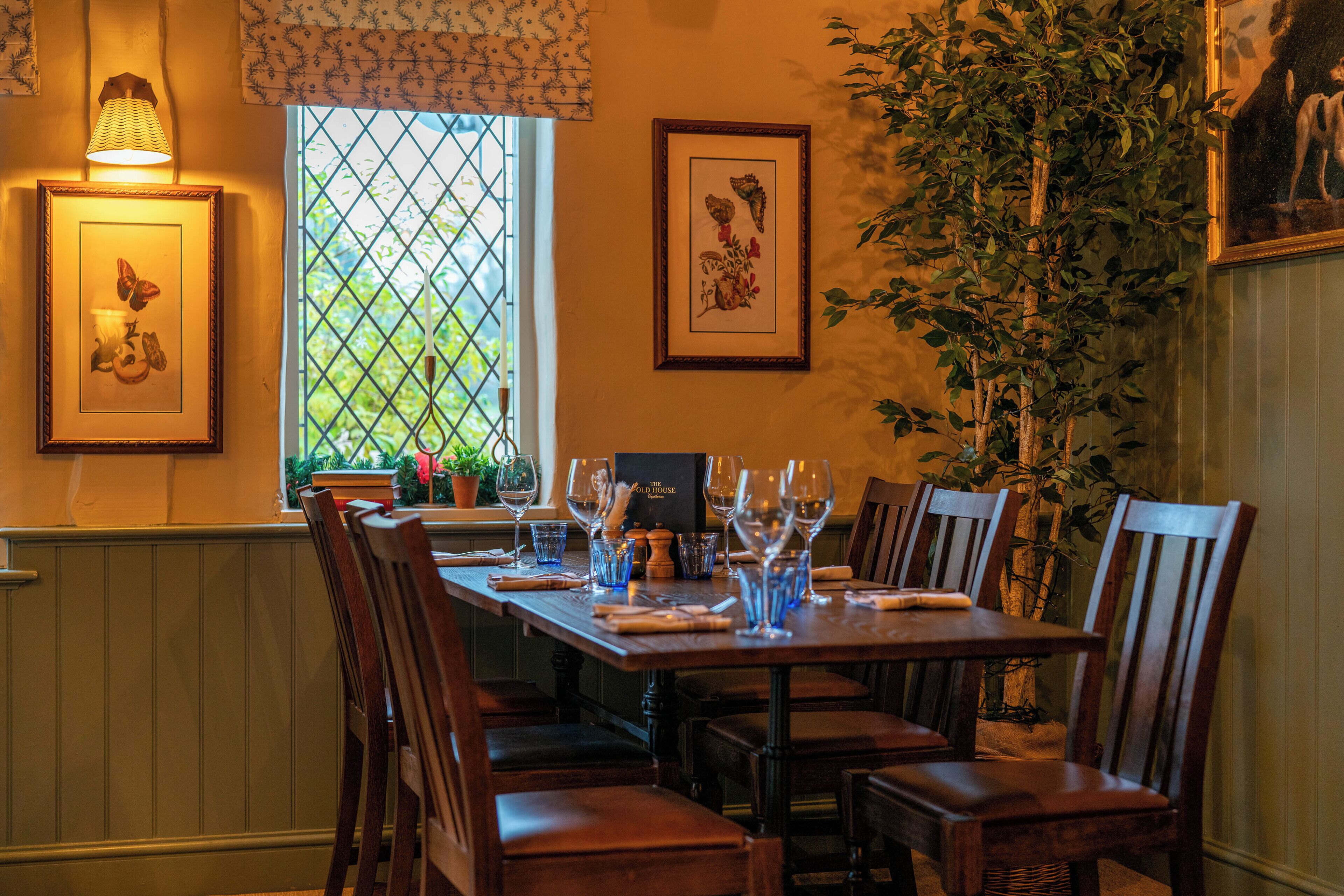 Un comedor con una mesa de madera dispuesta para seis personas, rodeada de sillas de madera. Se observan cuadros en las paredes, una ventana con vista a la vegetación y una planta en maceta al lado.