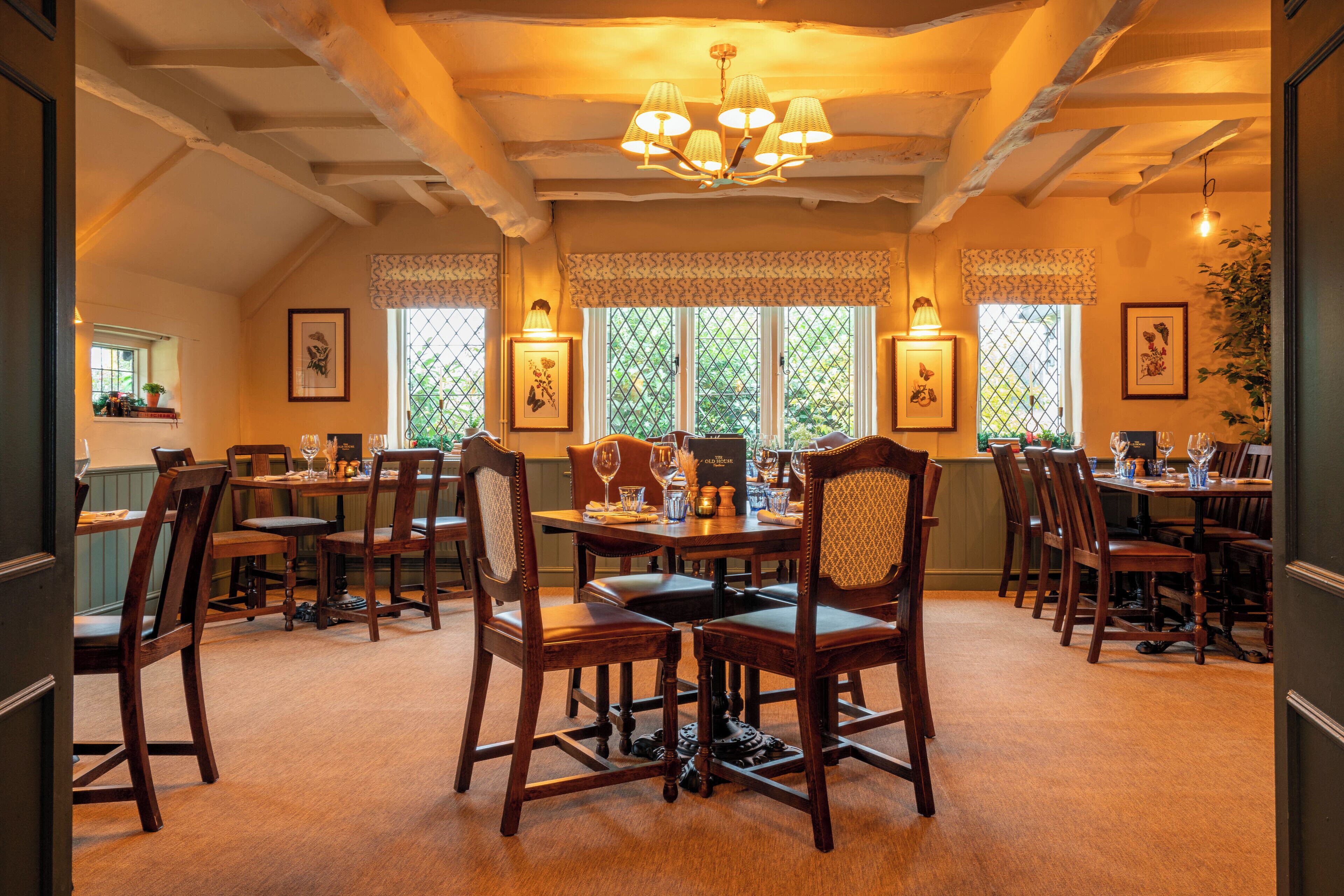 A dining area with wooden tables and chairs, a chandelier, and windows with patterned curtains.