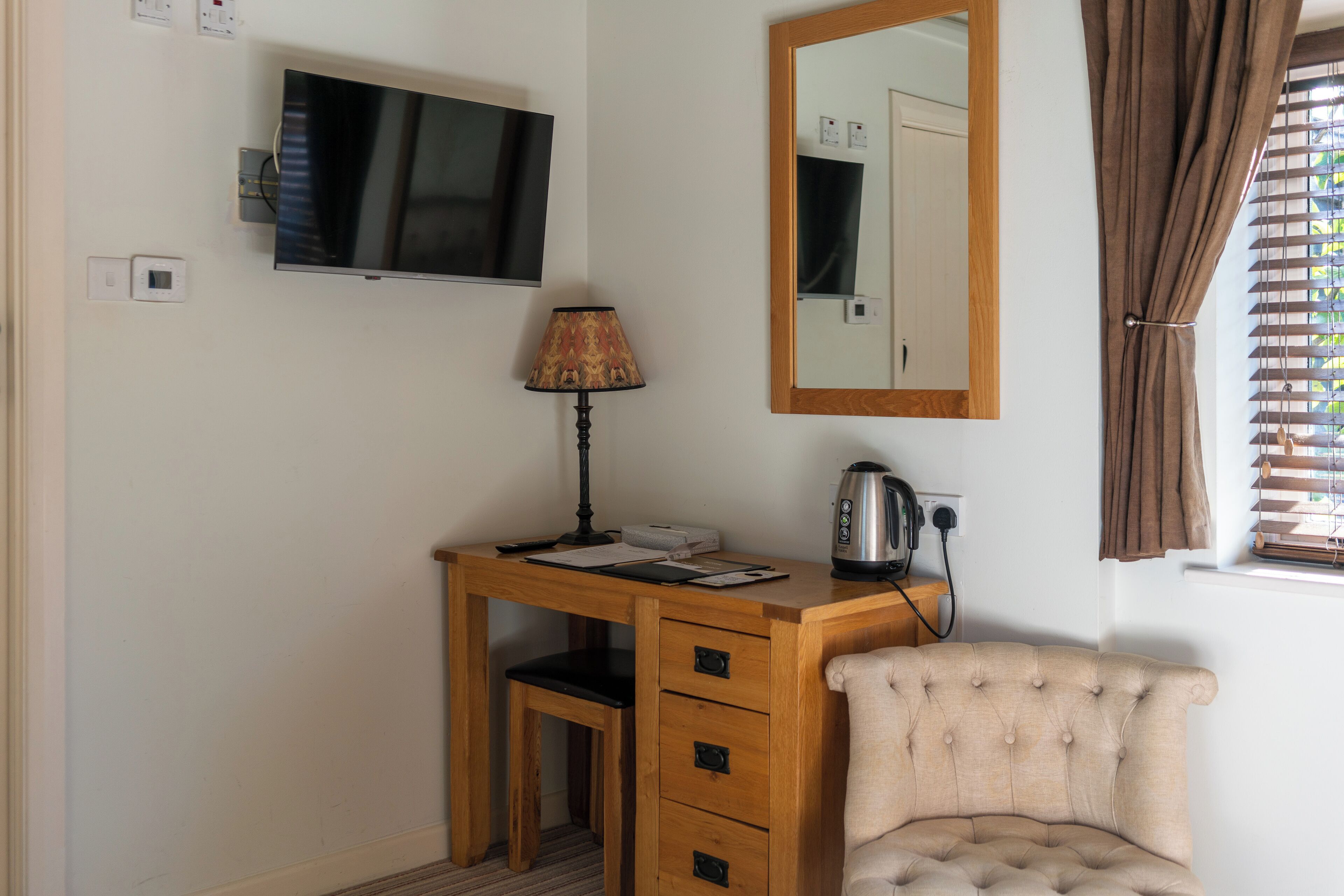 A wooden desk with a television, lamp, and kettle, a chair, and a mirror.