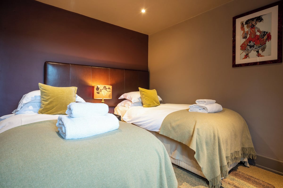Quarto de hotel com duas camas, cada uma com edredom branco e travesseiro amarelo. Há uma obra de arte emoldurada na parede e uma luminária na mesa de cabeceira.