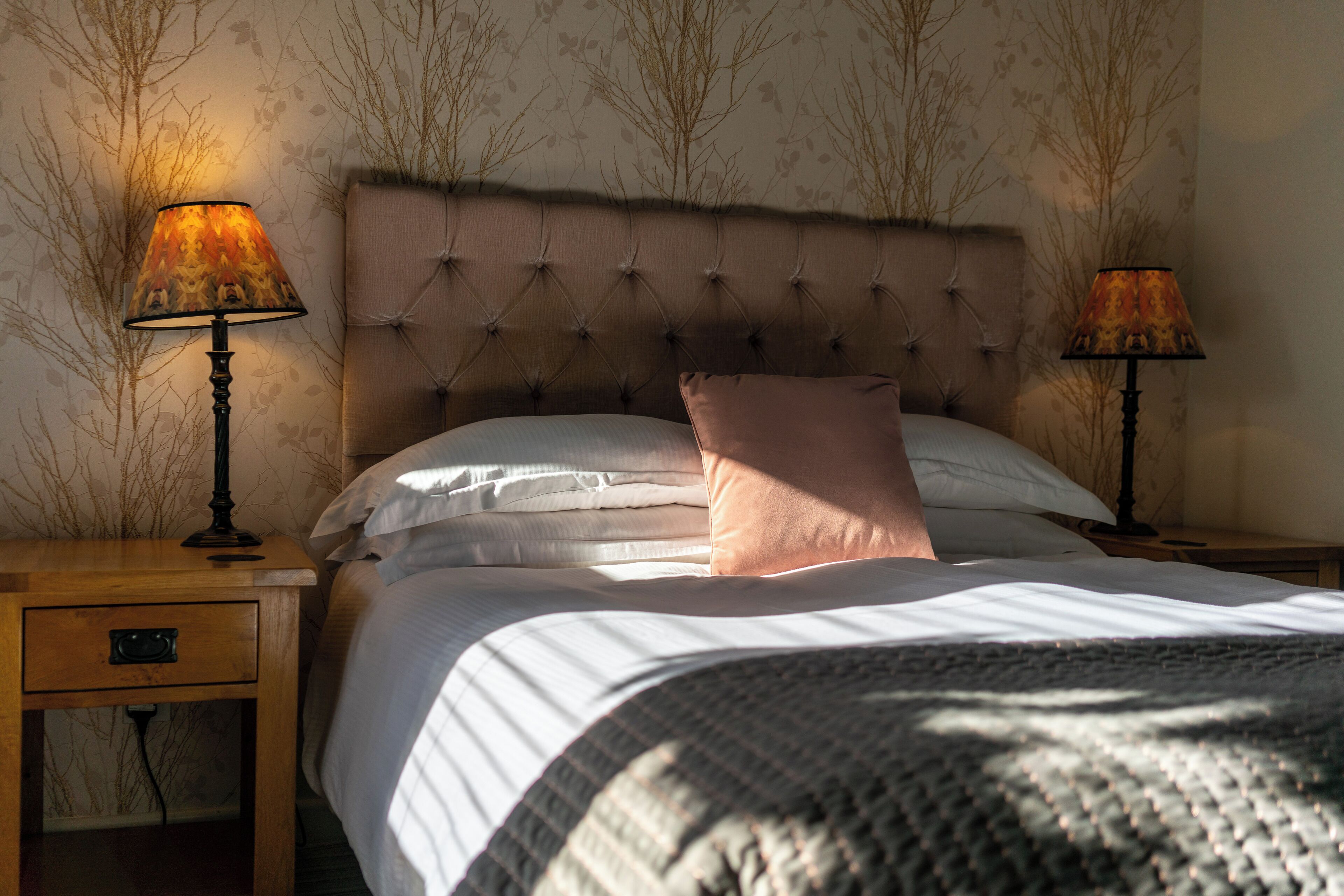 A neatly made bed with a tufted headboard, two bedside lamps, and a small wooden nightstand with a drawer.