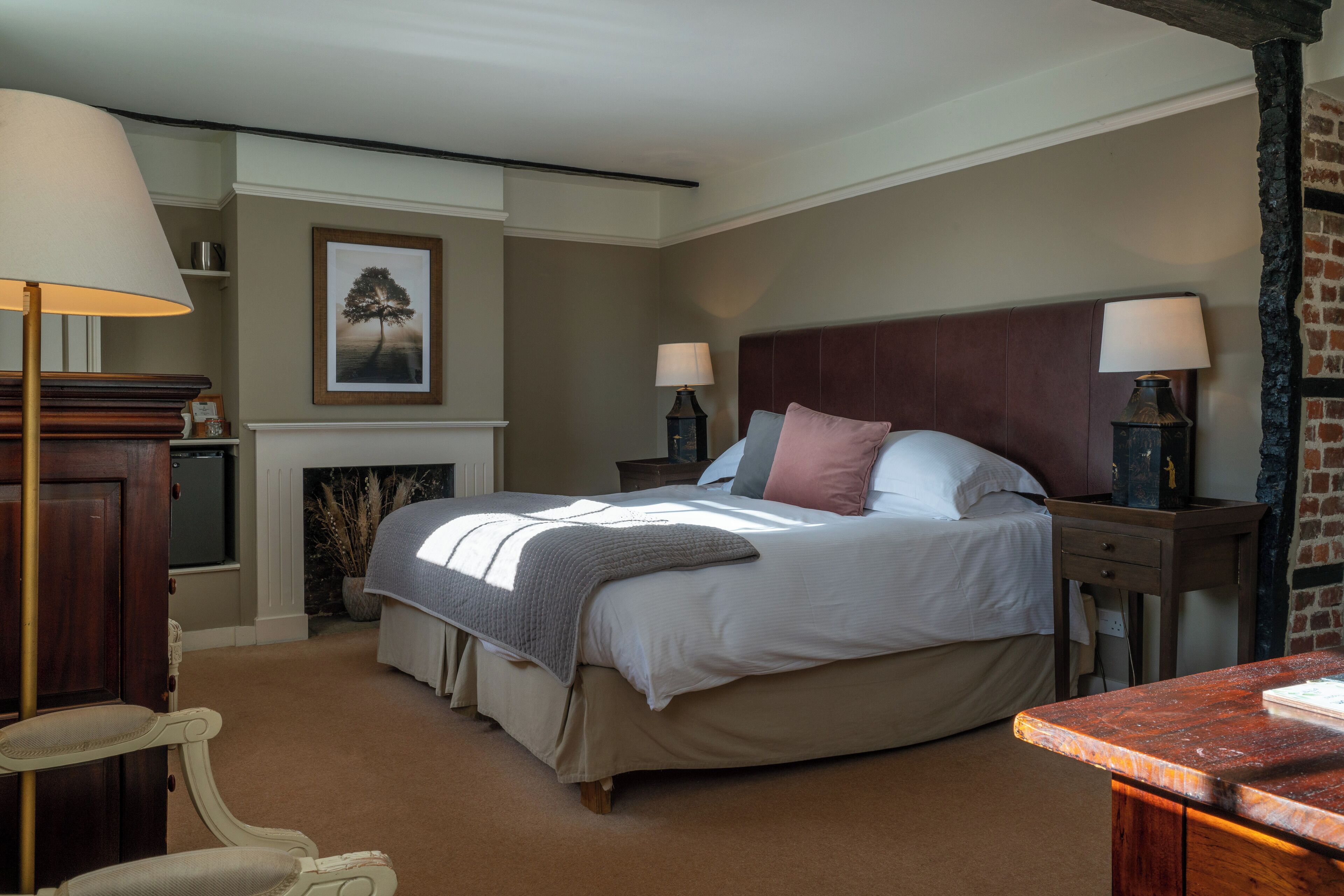 A bedroom with a large bed, two bedside tables with lamps, a chair, a fireplace, and a framed picture on the wall.