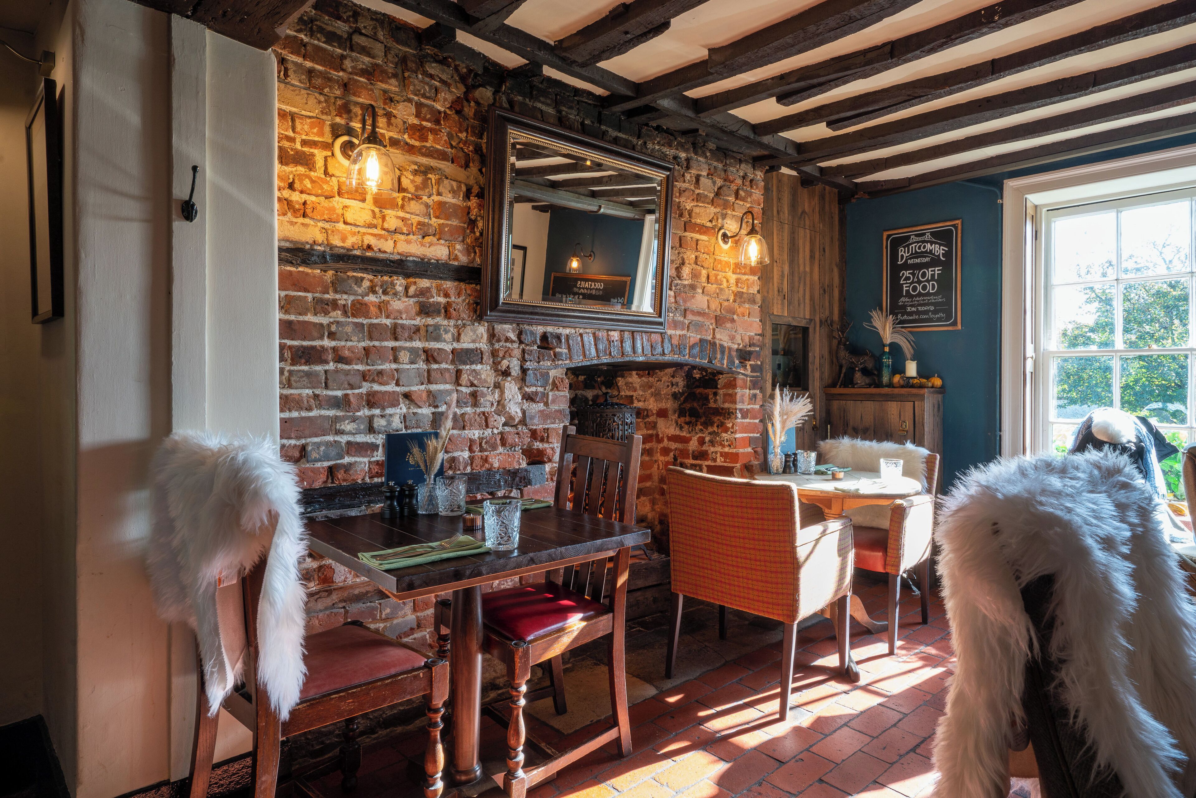 A rustic dining area with exposed brick walls, wooden beams, and a brick fireplace. There are two white chairs with fur trim, a wooden table, and a mirror above the fireplace. A chalkboard sign is visible on the wall.