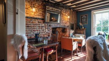 A rustic dining area with exposed brick walls, wooden beams, and a brick fireplace. There are two white chairs with fur trim, a wooden table, and a mirror above the fireplace. A chalkboard sign is visible on the wall.