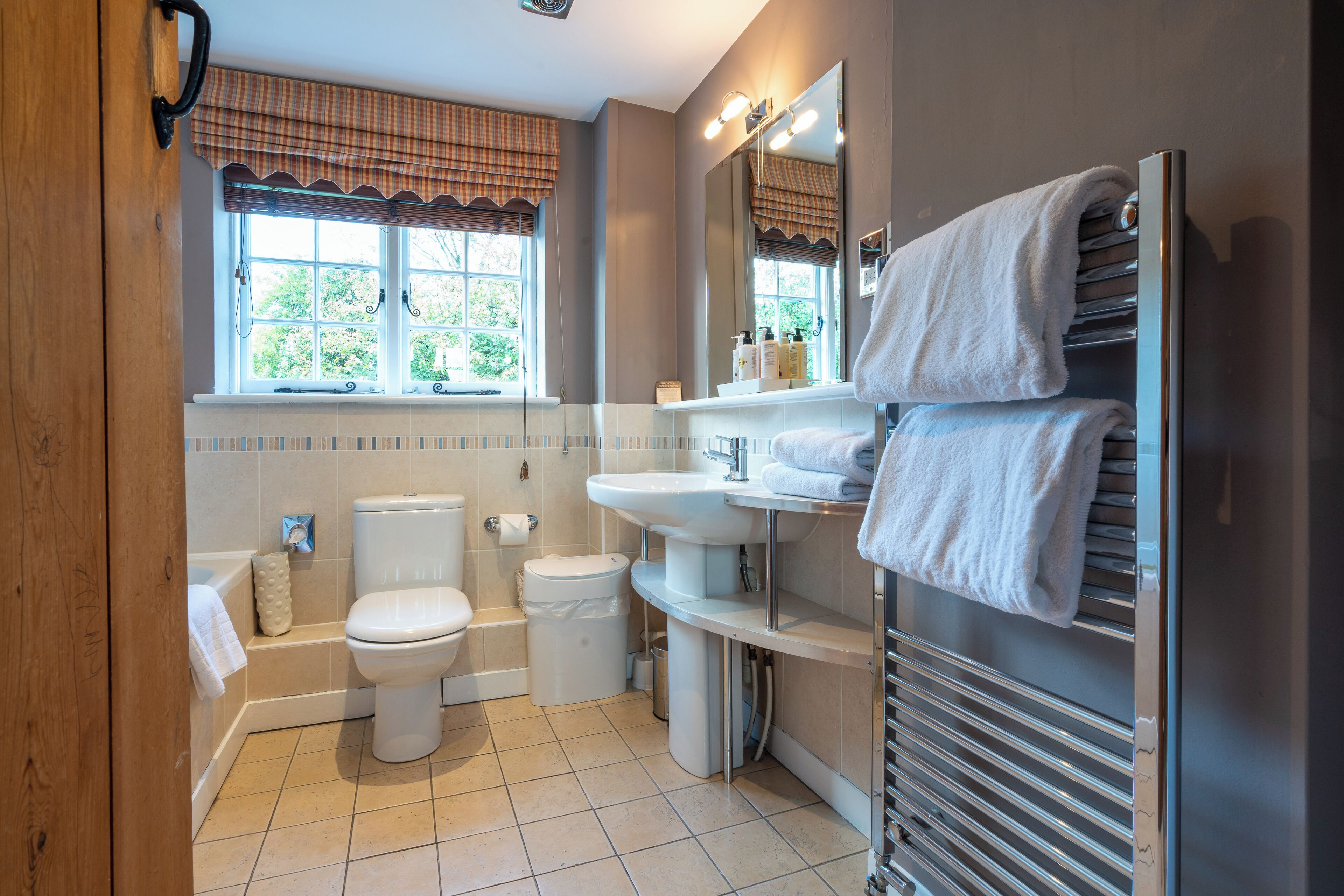 Een moderne badkamer met toilet, wastafel en bad. Er is een raam met gordijnen en een handdoekrek met handdoeken.