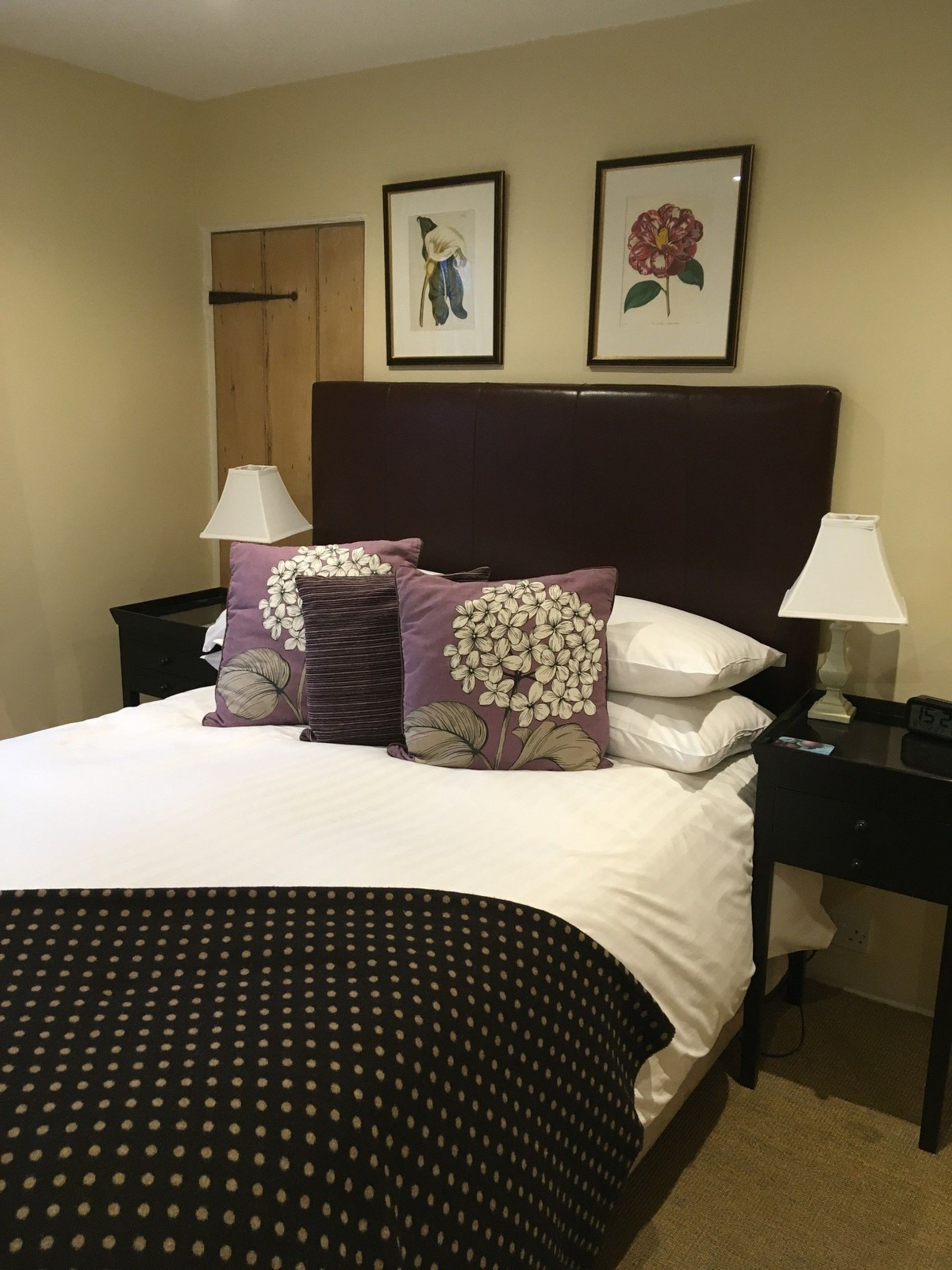 Una cama bien hecha con una colcha de lunares oscuros, dos almohadas moradas a juego con diseños florales blancos y un cabecero marrón oscuro. Encima de la cama, hay dos cuadros en la pared, uno con una persona y el otro con una flor.