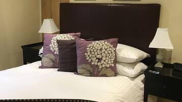 Una cama bien hecha con una colcha de lunares oscuros, dos almohadas moradas a juego con diseños florales blancos y un cabecero marrón oscuro. Encima de la cama, hay dos cuadros en la pared, uno con una persona y el otro con una flor.