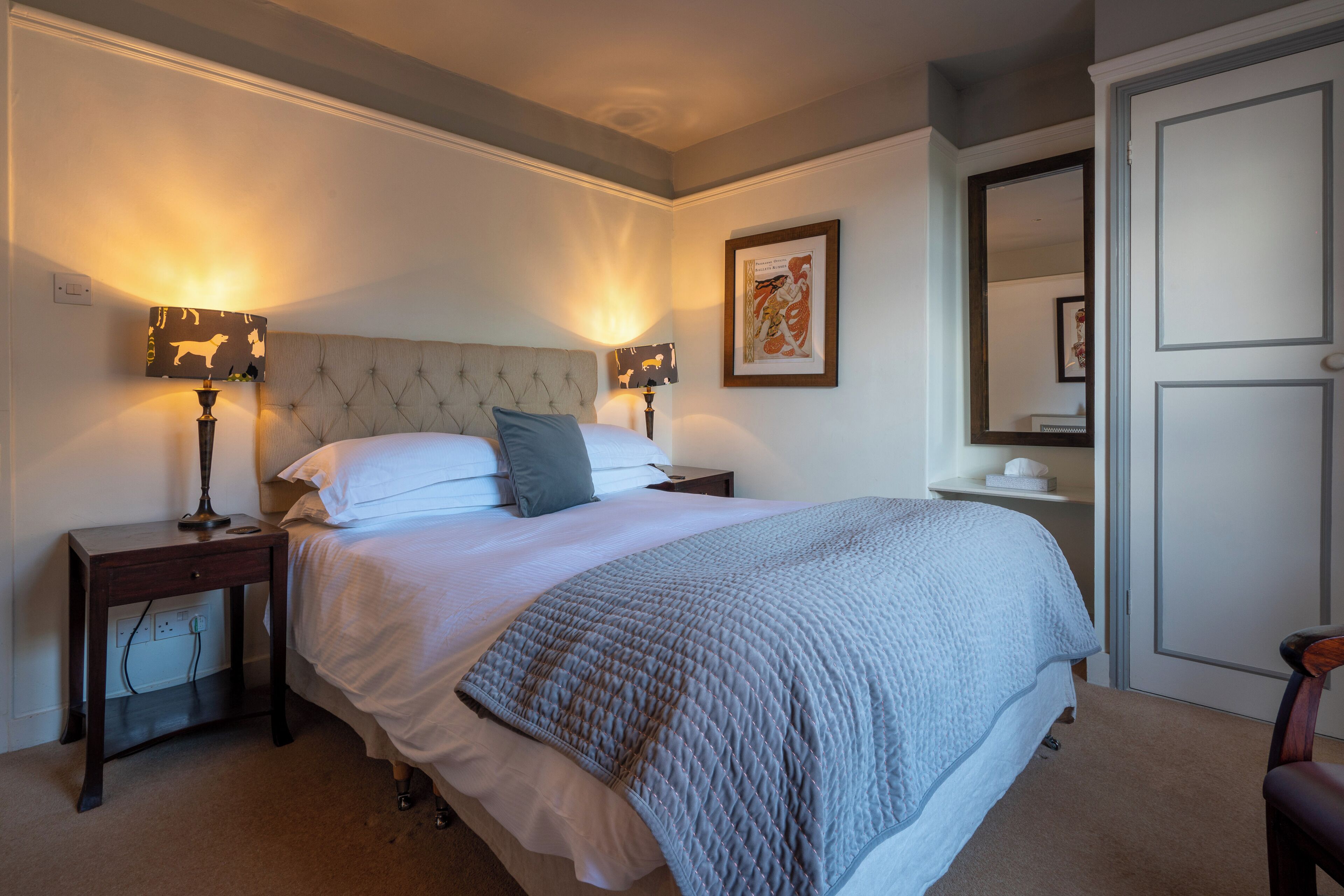A neatly arranged bedroom with a large bed, two bedside tables with lamps, a framed picture on the wall, and a door to the right.