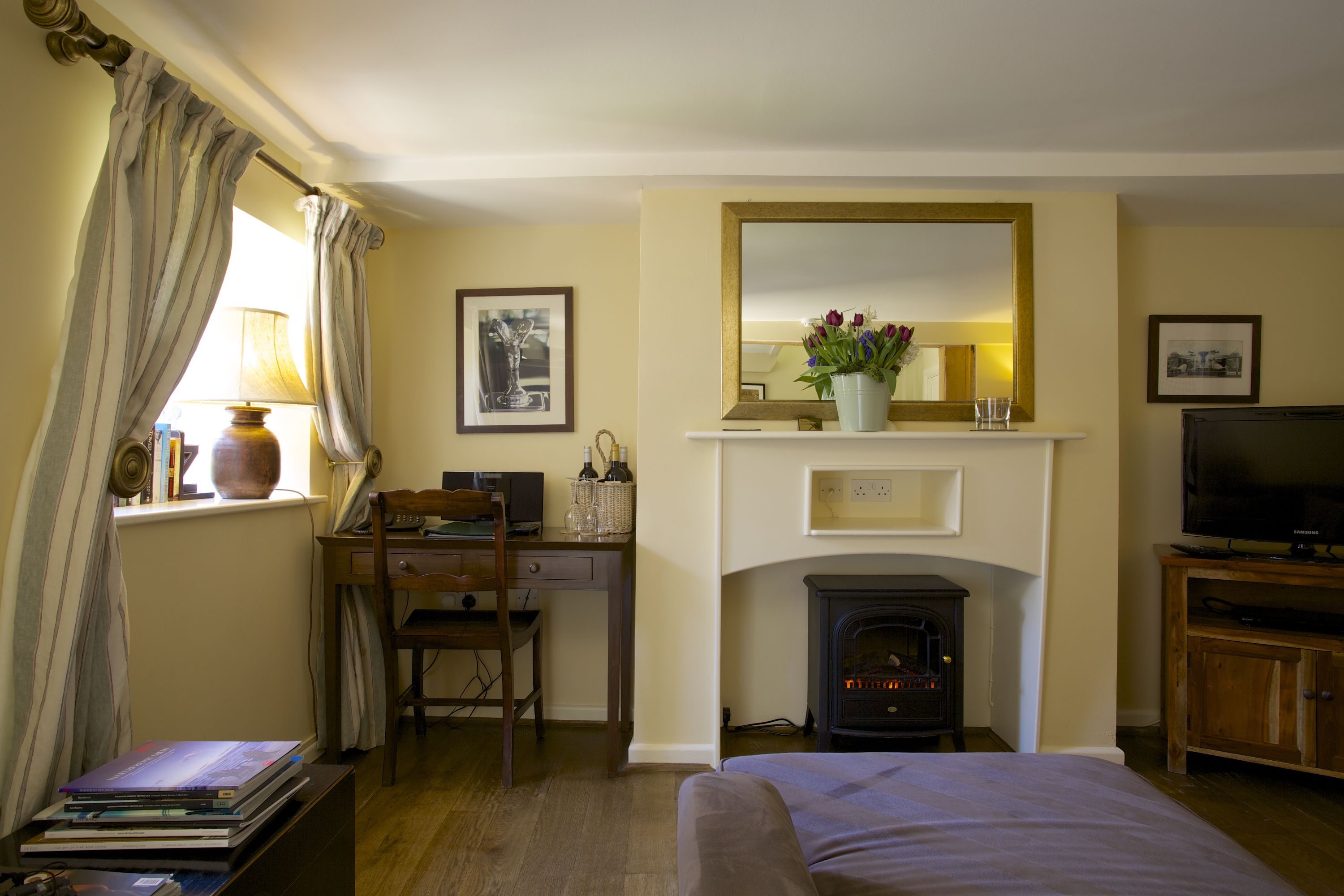 A hotel room with a bed, a desk with a chair, a television, a fireplace, and a mirror.