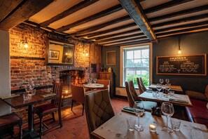 A cozy restaurant with brick walls, wooden beams, and a menu board on the wall. Tables are set with glasses and cutlery. - The Royal Oak (Chichester)