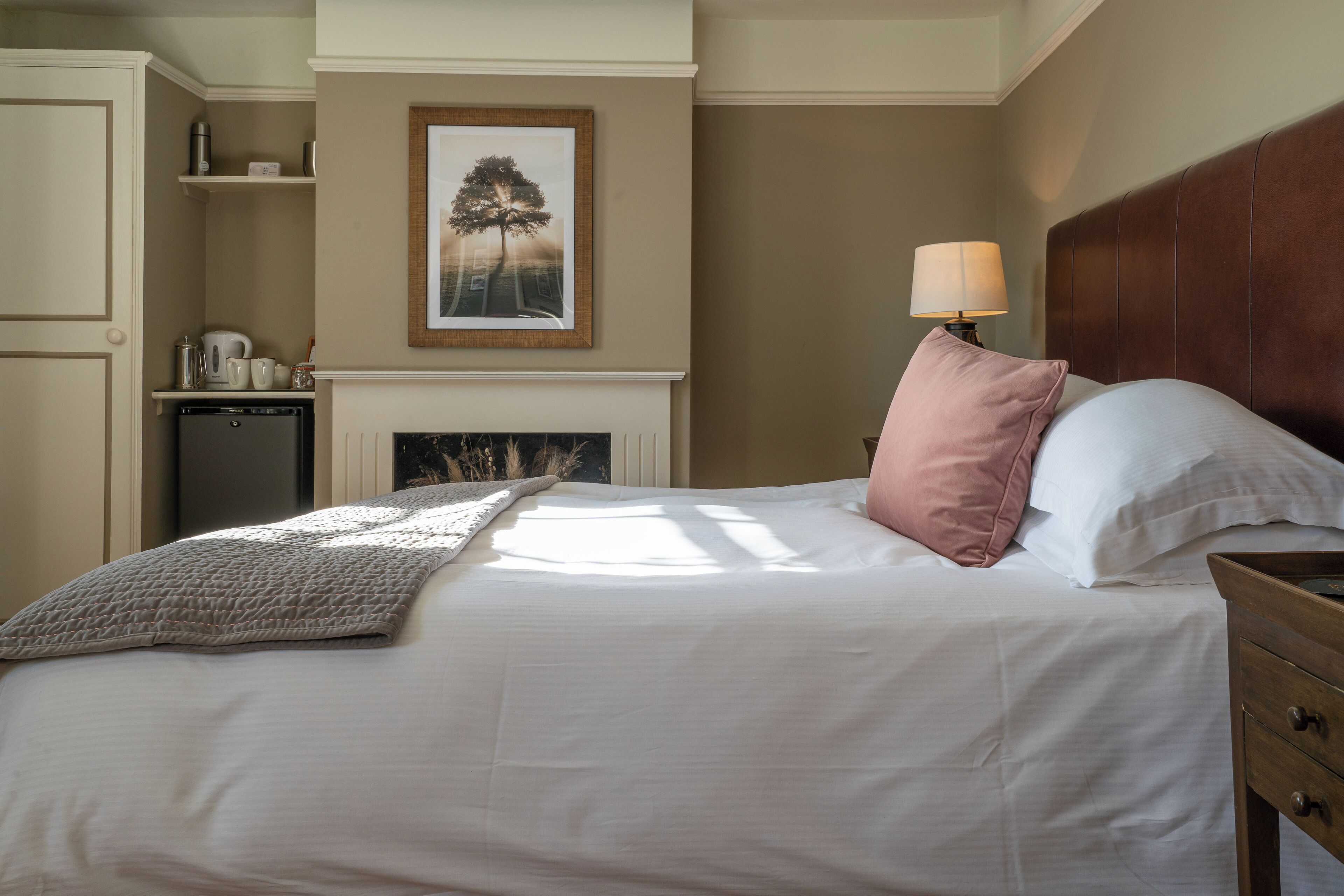 A neatly made bed with a white comforter and a pink pillow, a wooden nightstand with a lamp, a framed picture above the bed, and a small kitchen area with a coffee maker and kettle.