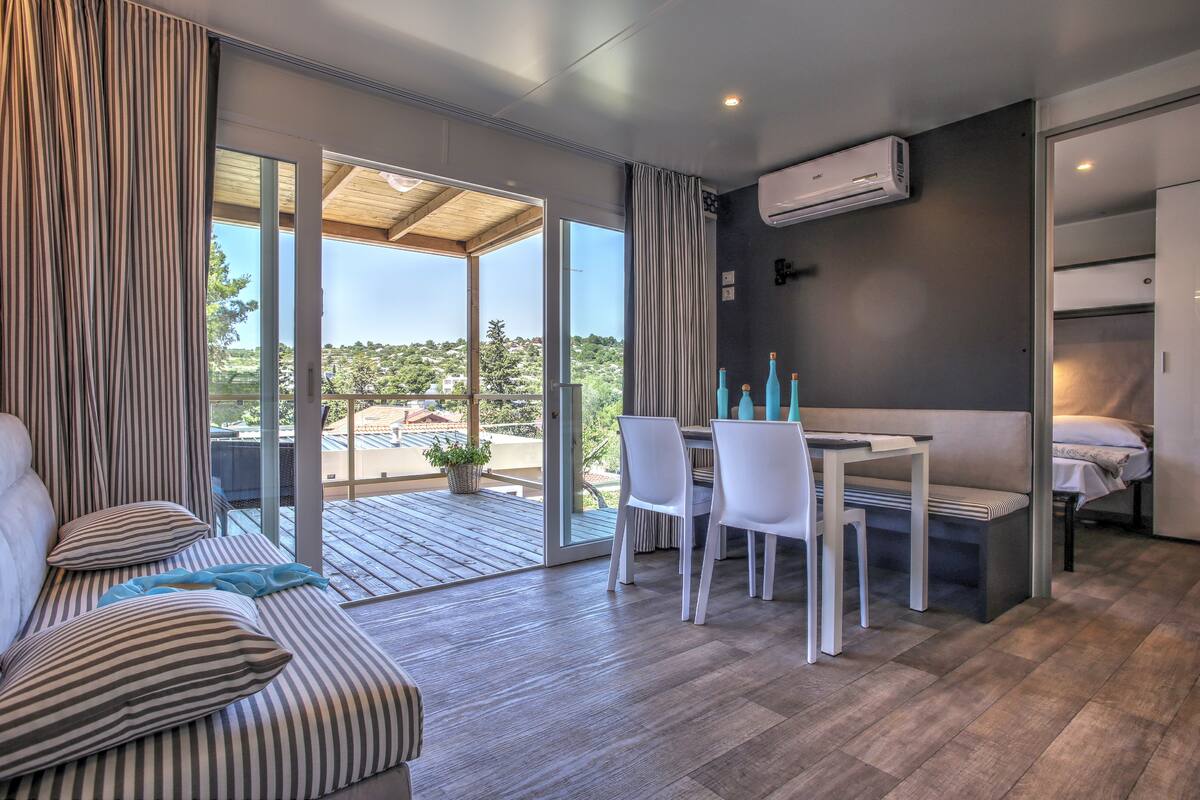 Quarto moderno com sofá, mesa de jantar com cadeiras, cama pequena e porta de vidro deslizante para área externa.