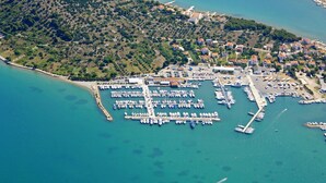 An aerial view of a coastal marina with numerous boats docked, a pier extending into the water, and a town with buildings and greenery in the background. - Camp Rehut - Campsite (Murter-Kornati)