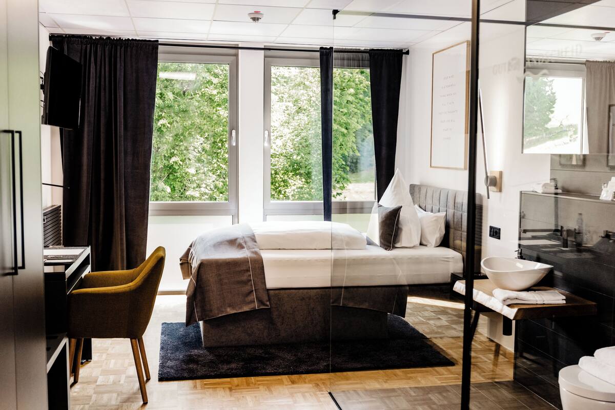Una habitación de hotel moderna con cama, escritorio, silla, ventana grande y baño.