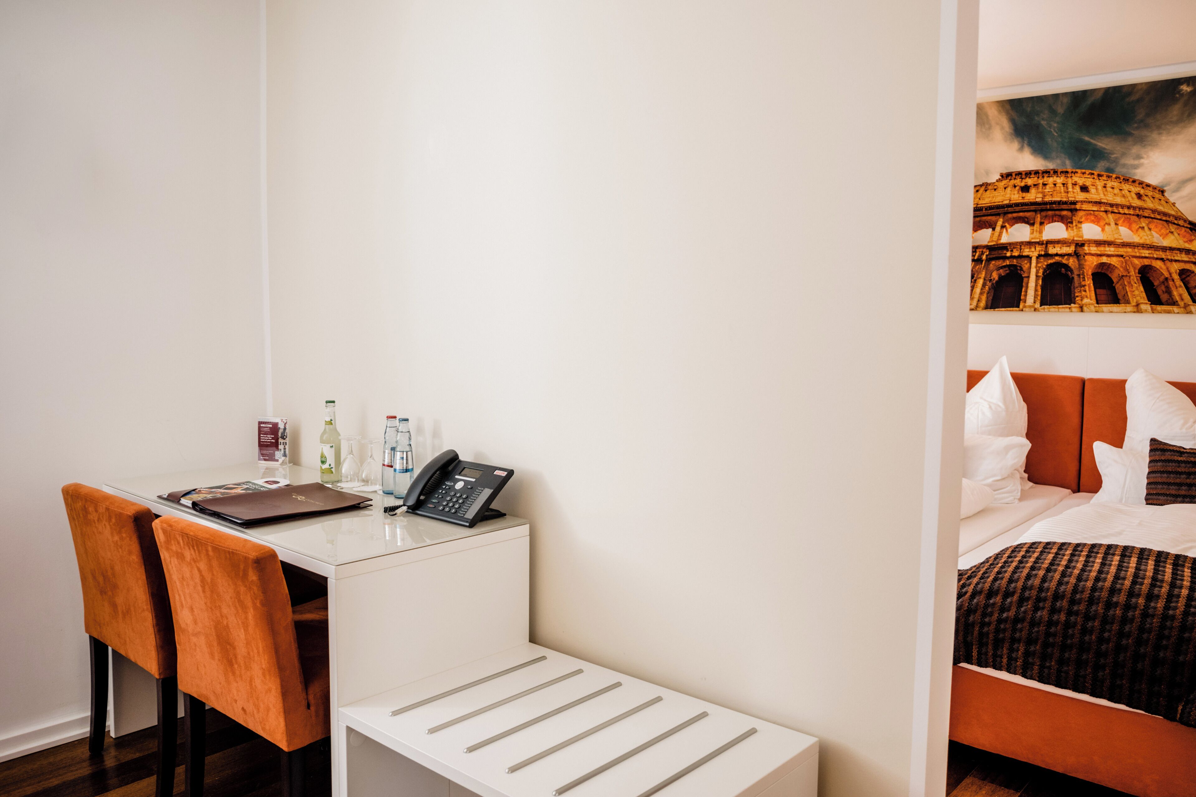 Een hotelkamer met een wit bureau, twee oranje stoelen, een bed met oranje hoofdeinde en een muurschildering van het Colosseum.