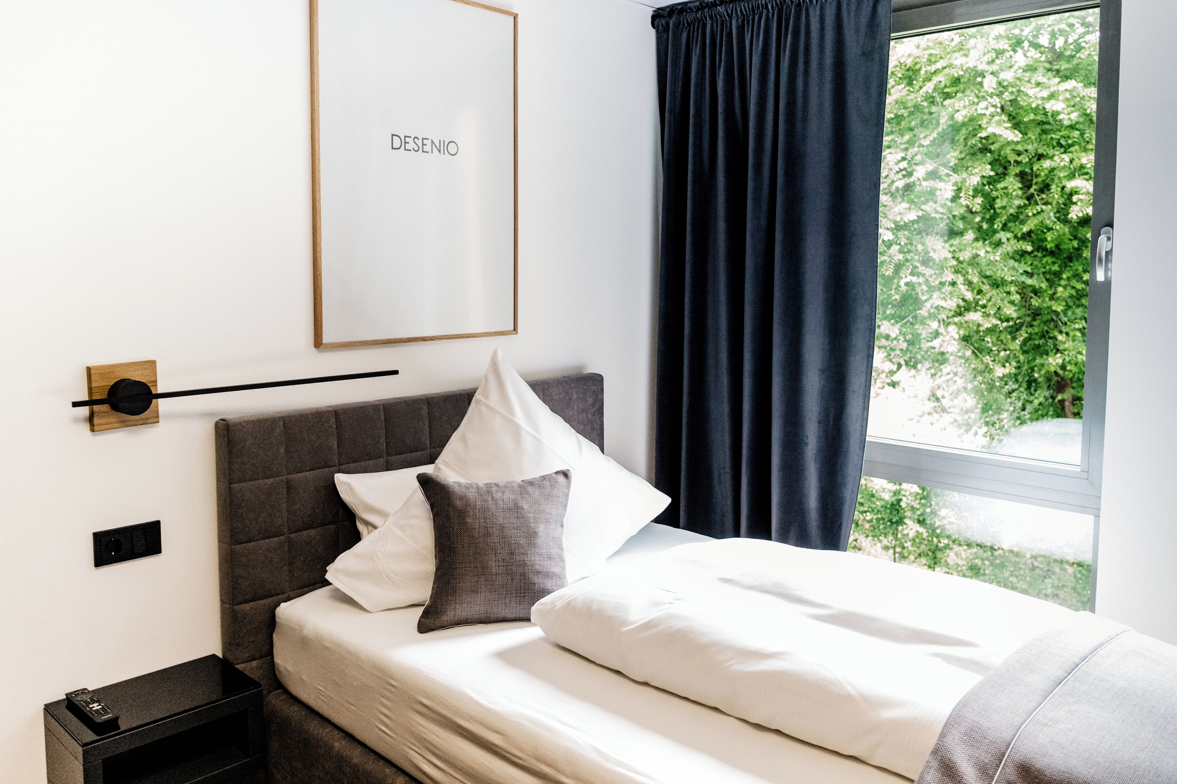 Ein ordentlich bezogenes Bett mit weißer und grauer Bettwäsche, ein gerahmtes Bild an der Wand und ein Fenster mit Blick auf Grünflächen.