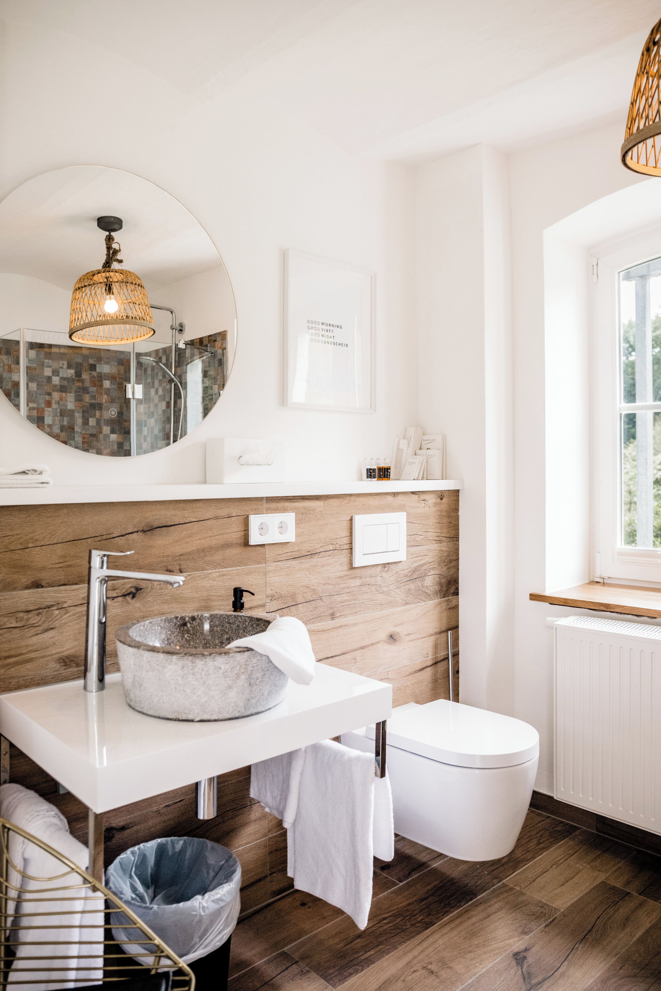 A modern bathroom with a round mirror, a stone sink, a toilet, and a window.