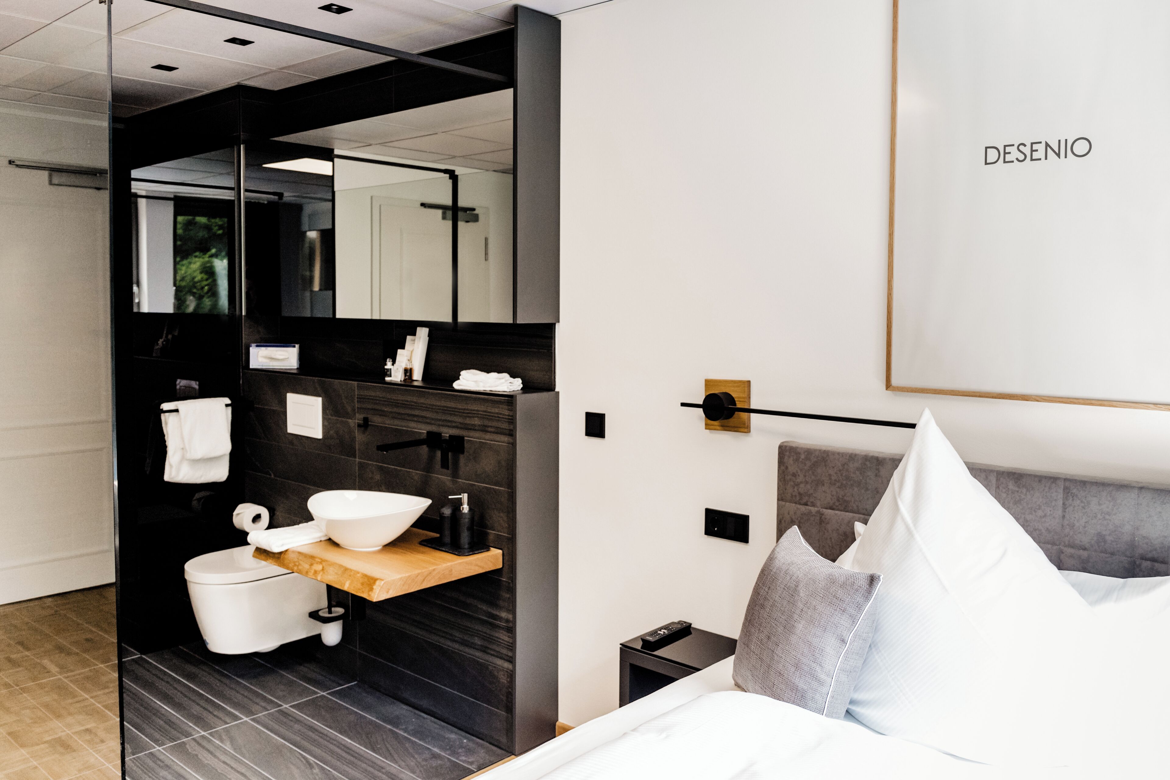 A modern hotel room with a large bed, a wooden bedside table, a built-in sink, and a mirror.