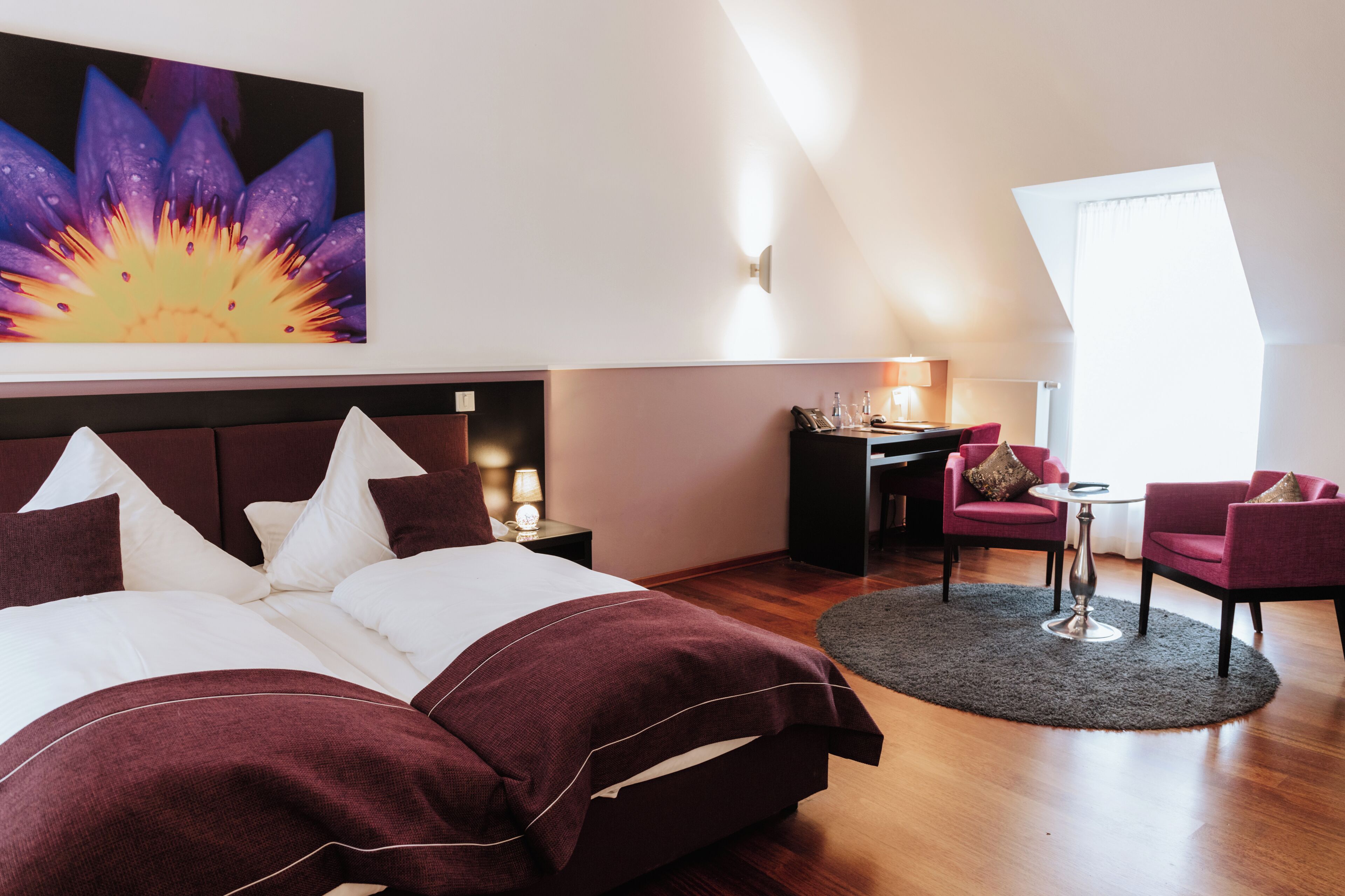 A modern hotel room with a large bed, a desk, two chairs, and a large window.