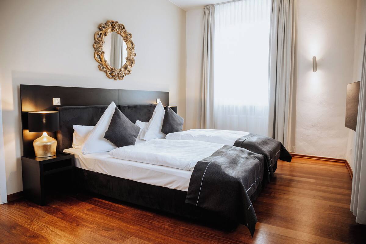 Habitación de hotel con cama doble, mesita de noche con lámpara, espejo y ventana con cortinas.
