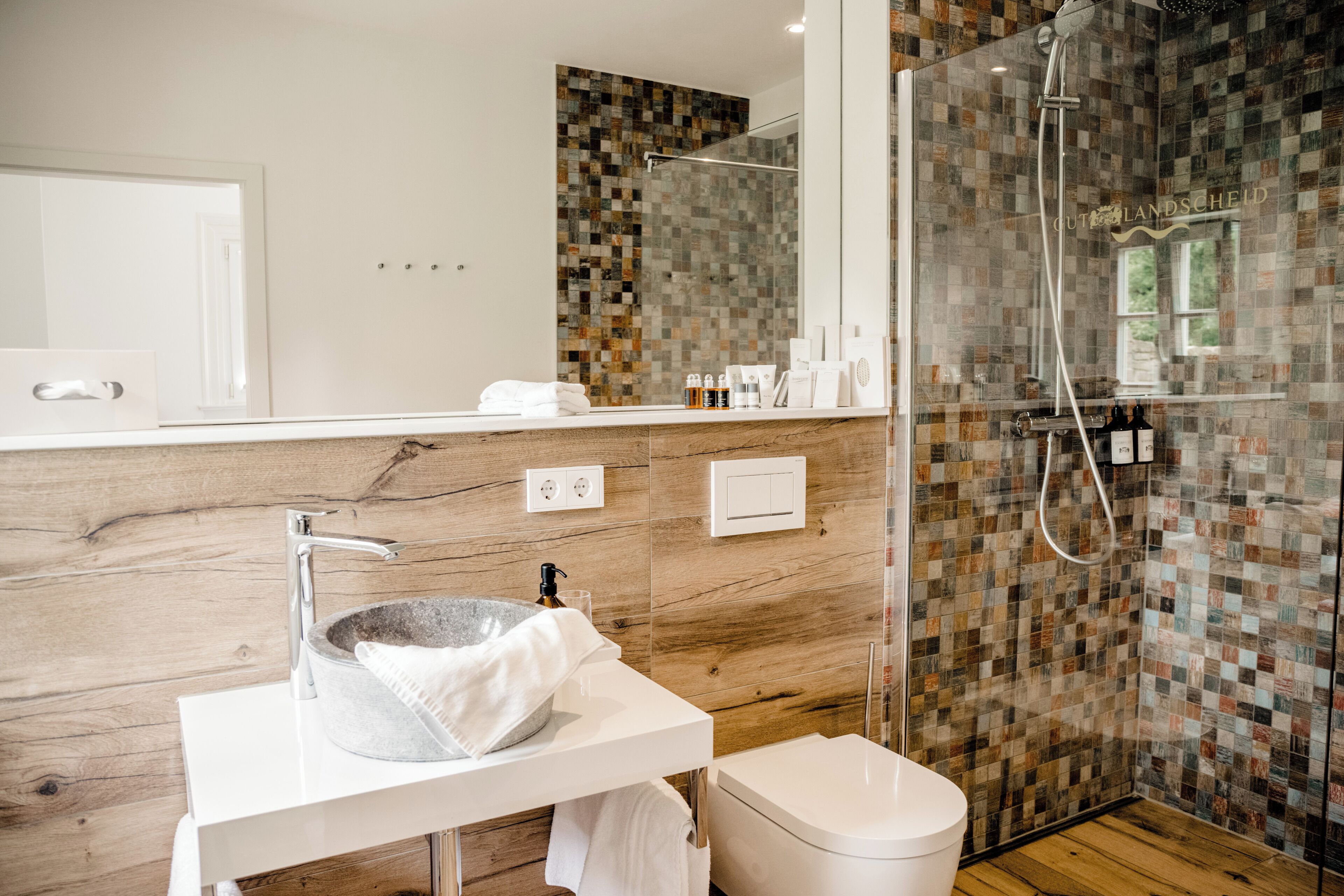 A modern bathroom with a stone sink, white fixtures, and a mosaic shower wall.