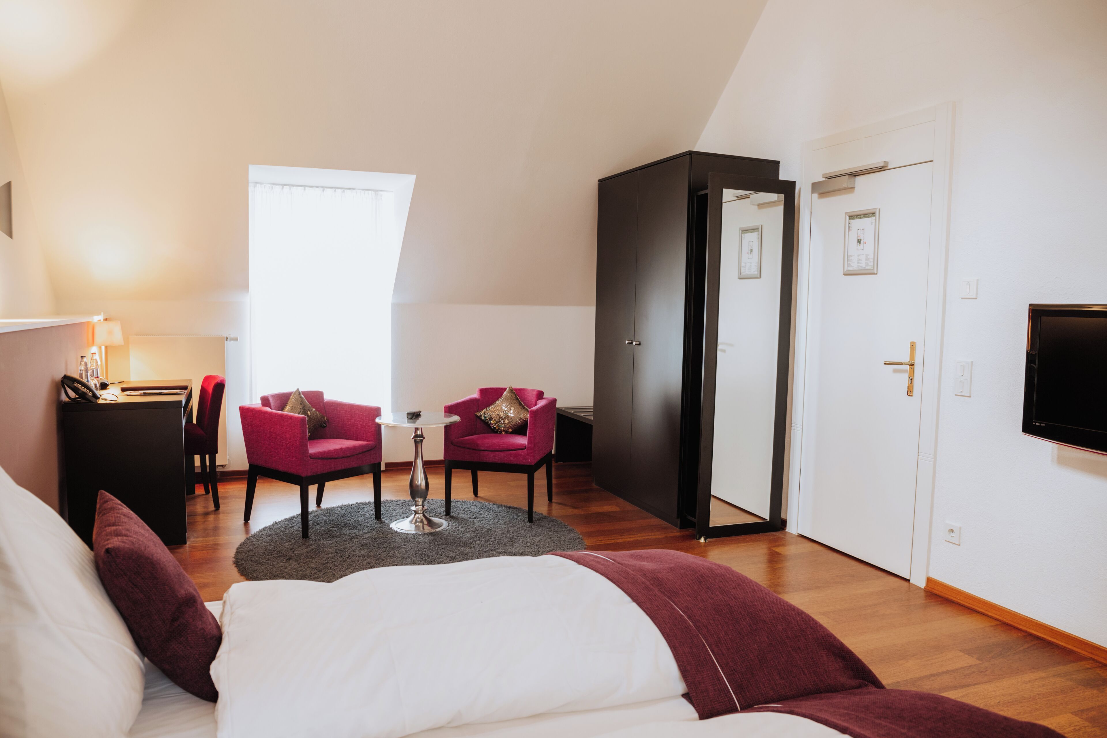 A modern hotel room with a bed, a desk, two chairs, a TV, and a door.