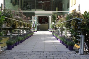 Entrada de hotel com fachada de vidro, escadaria metálica e plantas em vasos ao longo do caminho.