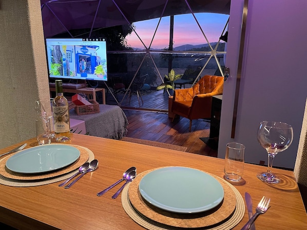 A dining area with a table set for two, a bottle of wine, and a television displaying a room. - Shambala Piri (Pirenópolis)